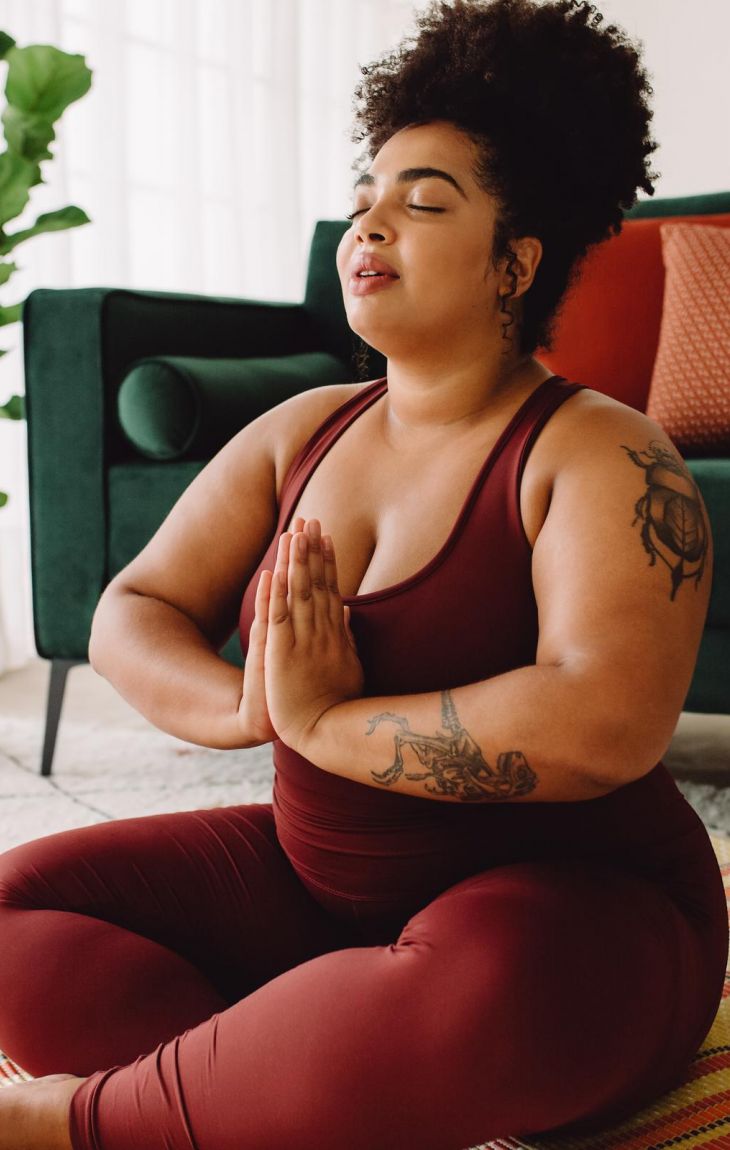 Woman in maroon activewear meditating cross-legged on a yoga mat in a cozy living room.