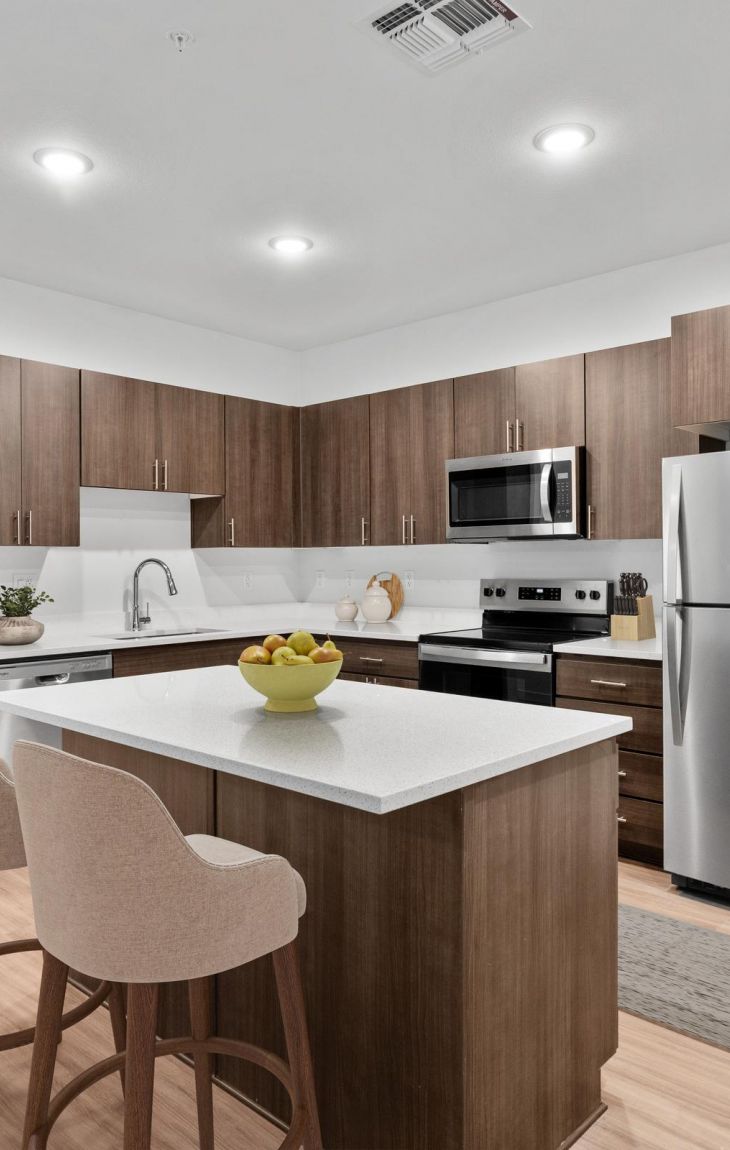 Modern kitchen with wood cabinets, island, two chairs, stainless steel appliances, and bowl of fruit.