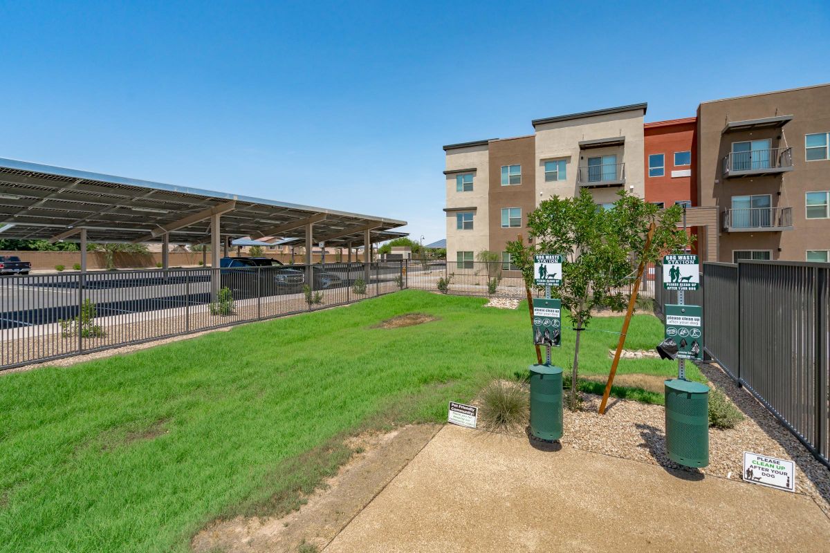 Fenced grassy dog park area with waste stations; apartments and covered parking in the background.