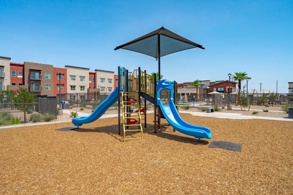 A playground with two blue slides, a climbing structure, and a sunshade, surrounded by wood chips and apartments.