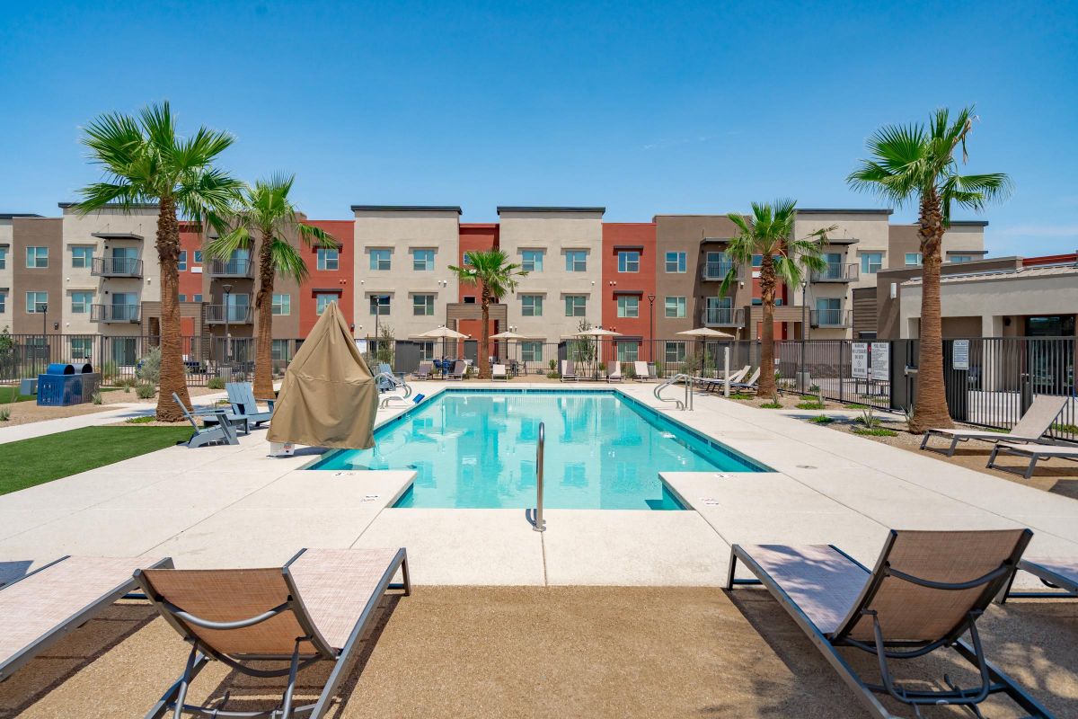 Modern apartment complex with a pool, lounge chairs, palm trees, and three-story buildings in the background.