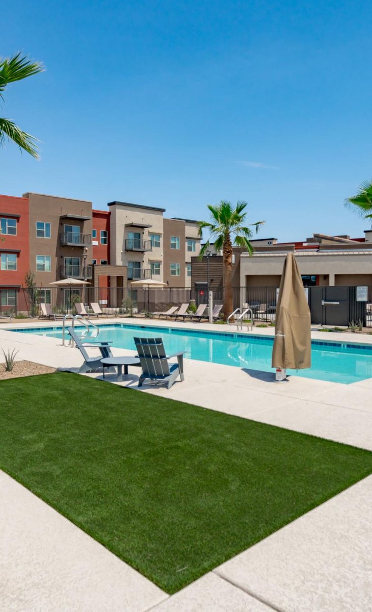 Modern apartment complex with palm trees, pool, lounge chairs, and clear blue sky.