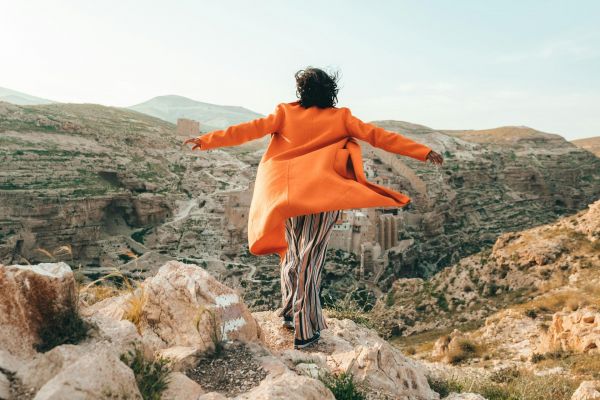 Person in an orange coat stands on rocky terrain with arms outstretched, overlooking a scenic canyon landscape.