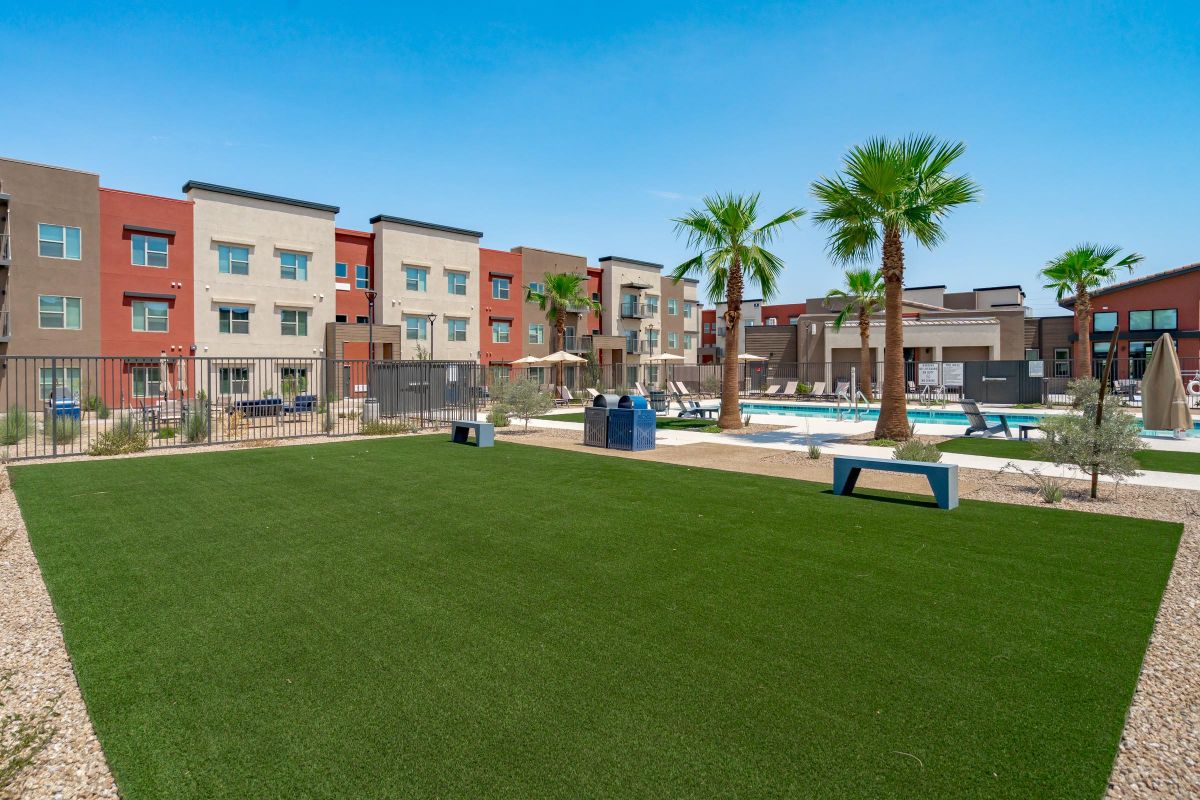 Fenced grassy courtyard with benches, palm trees, and a pool area in front of modern apartment buildings.