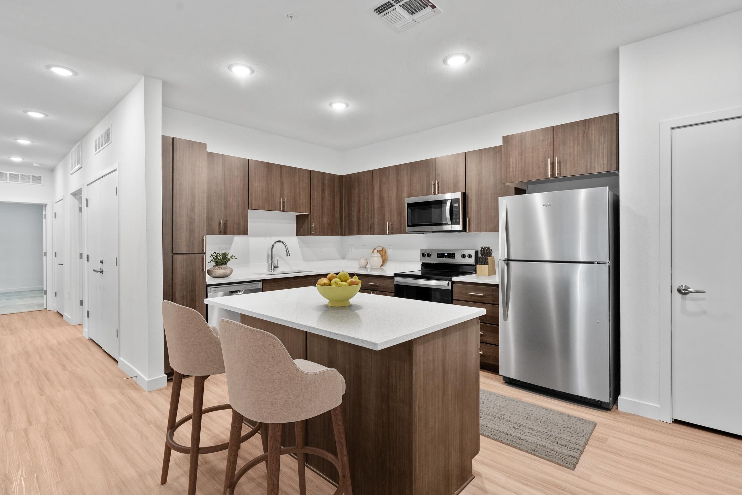 Modern kitchen with wood cabinets, island, two chairs, stainless steel appliances, and bowl of fruit.
