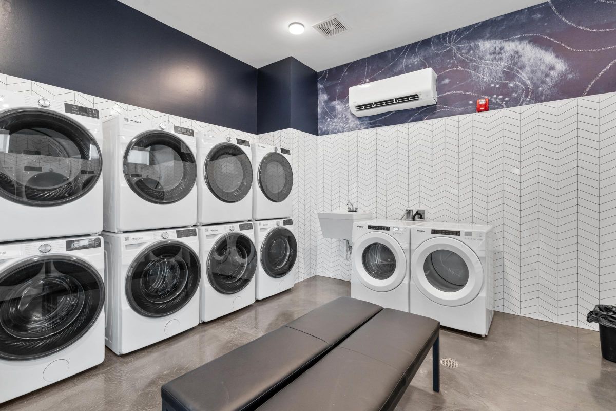 Modern laundry room with stacked washers and dryers, a black bench, and a sink against patterned tile walls.
