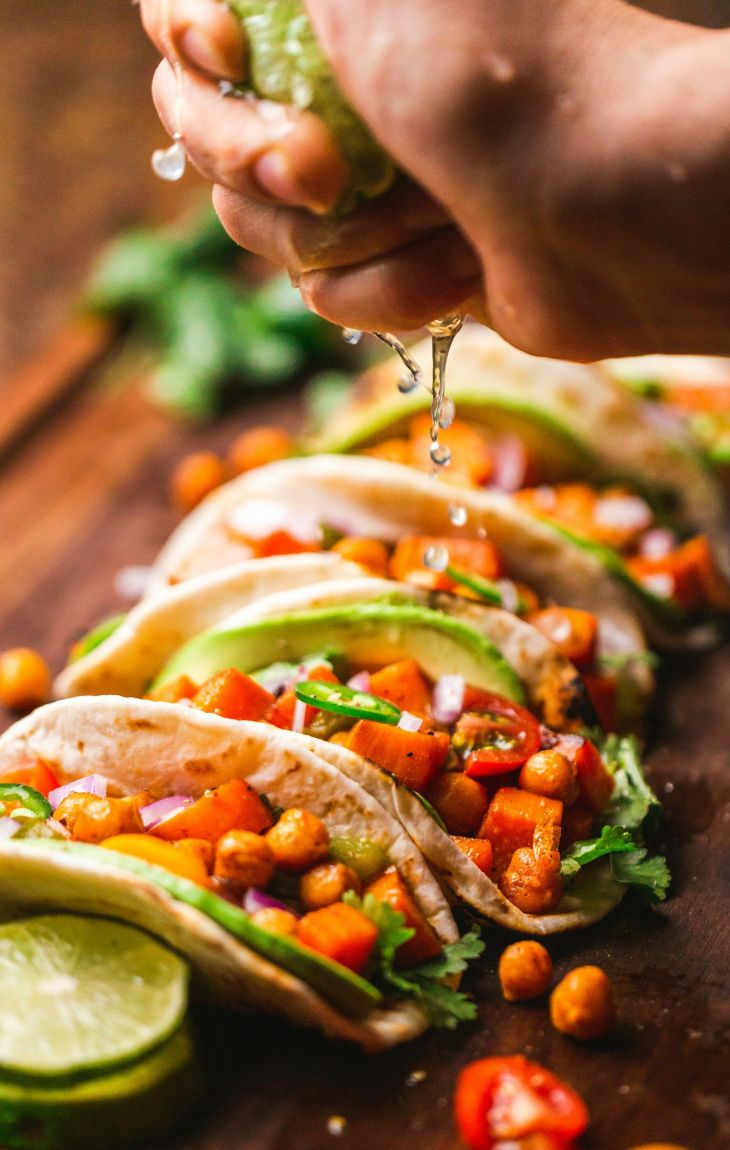 A hand squeezes lime over vegetable-filled tacos on a wooden board, garnished with fresh herbs.