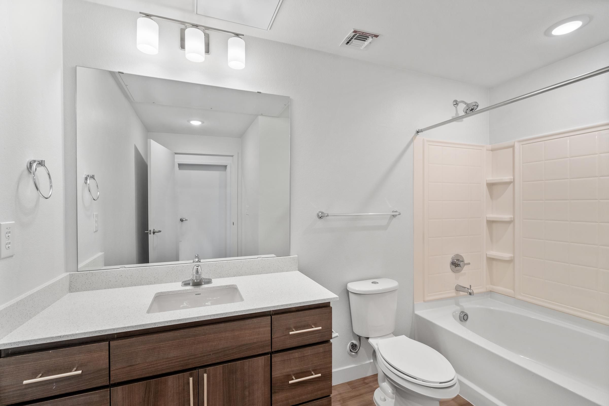 Modern bathroom with a wood vanity, large mirror, toilet, and bathtub with a beige shower surround.