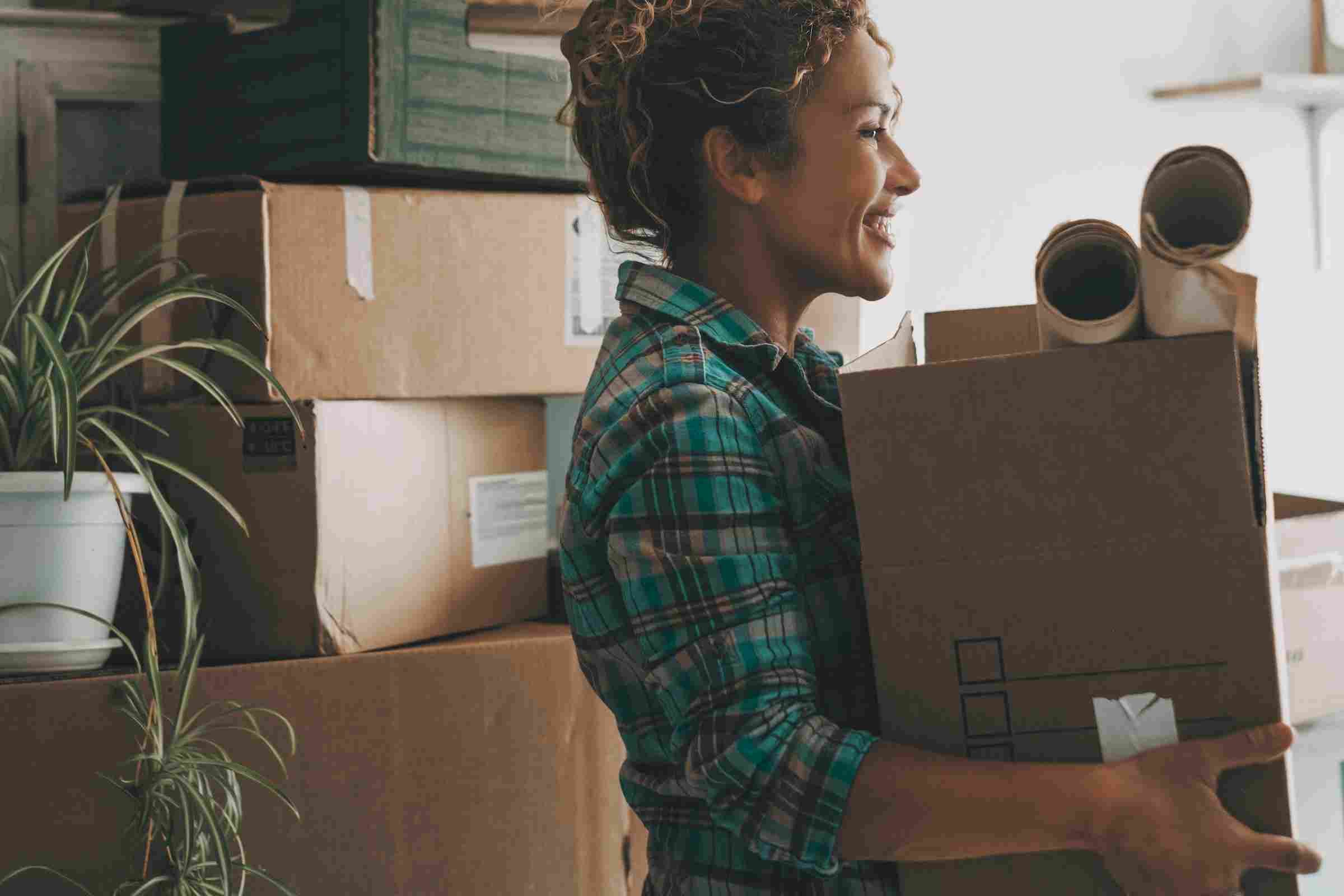 Affordable Apartments with Smiling woman holding a box and packing supplies, surrounded by moving boxes and a potted plant.