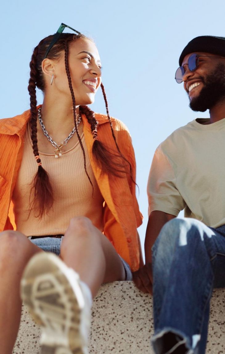 Two friends sit on a ledge outdoors, smiling at each other against a clear blue sky.