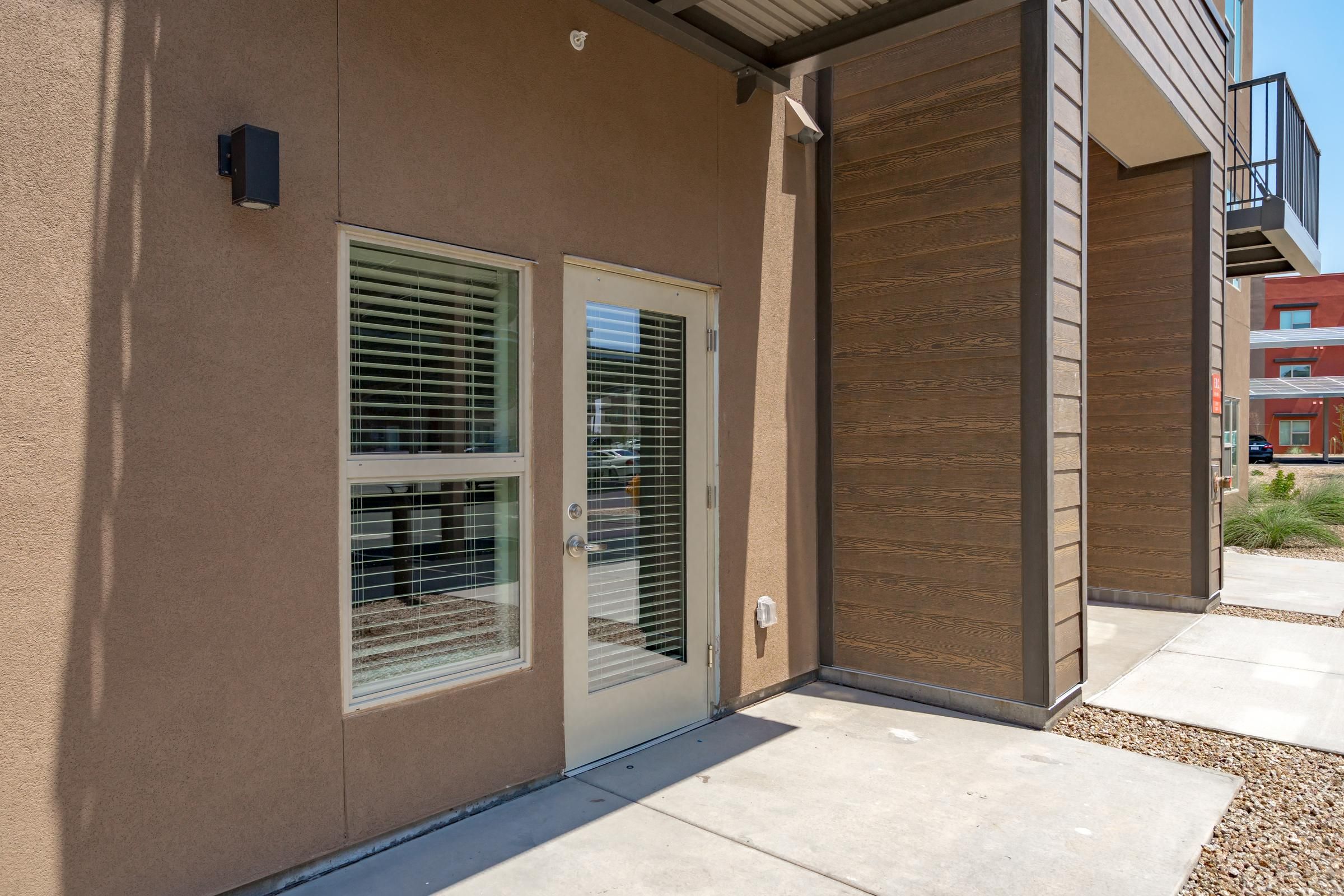 Glass door and window with white blinds on a modern brown building, with a concrete walkway in front.