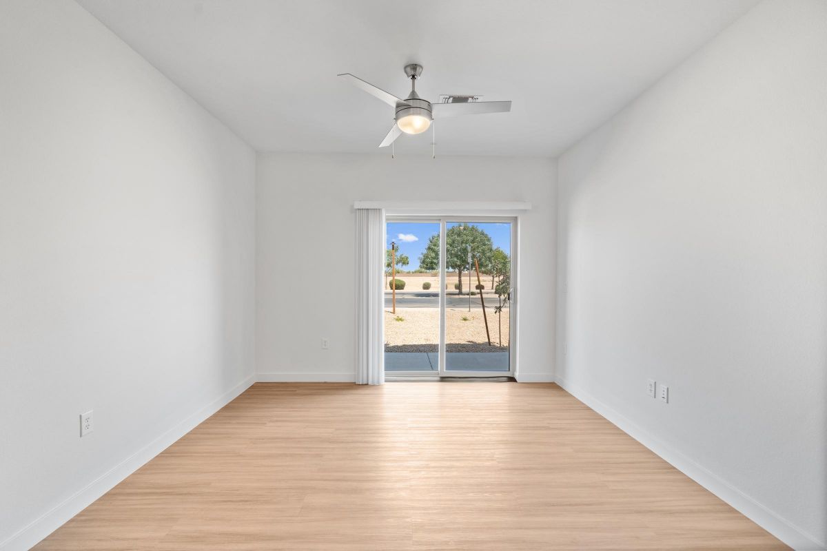 Empty room with light wood floor, white walls, ceiling fan, and sliding glass door opening to a patio with trees outside.